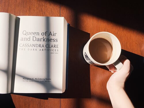 Queen of Air and Darkness by Cassandra Clare sitting open on a table with a hand and mug of coffee beside it.