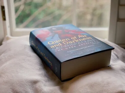 A hardback copy of Queen of Air and Darkness, Book Three of the Dark Artifices sitting on a pillow in front of an open window.